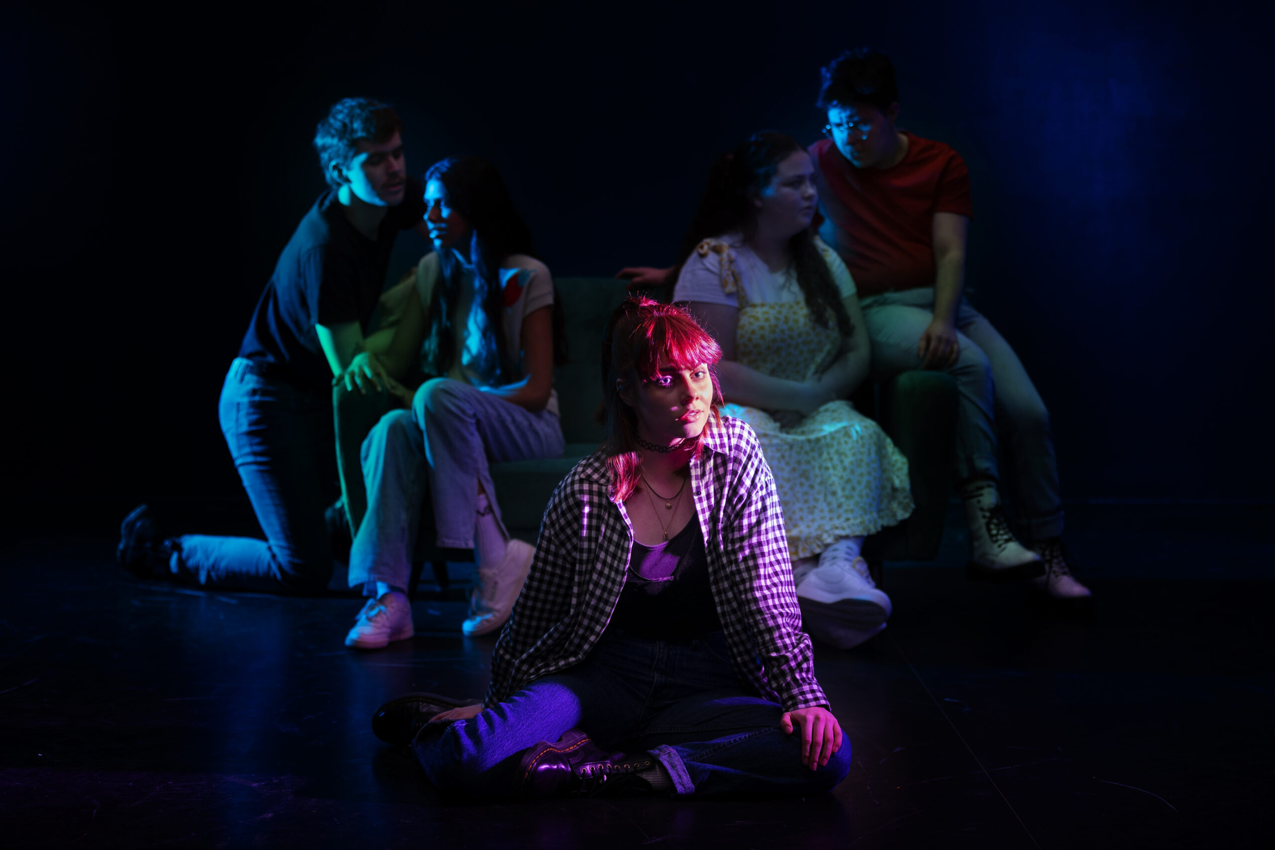 A young woman with pink hair sits on the floor and looks knowingly at the viewer. Two couples sit on a sofa behind her, talking together in semi-darkness.